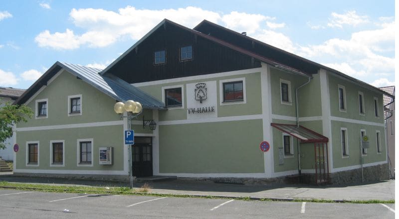 Die TV-Halle von außen: hellgrünes Haus mit weißen Lisenen und dem Vereinswappen über dem Schriftzug „TV-Halle“